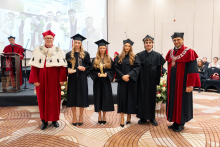 A group of people pose during an academic ceremony; three people in black academic gowns display statuettes, with people in red academic attire standing on either side.
