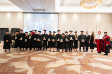 A group of people in black academic gowns and mortarboards pose for a group photo, displaying green statuettes; people in ceremonial academic attire stand on either side.