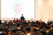 A view of the hall during an academic ceremony; a person in a black academic gown and mortarboard speaks from the lectern to a large audience, with the university emblem displayed in the background.