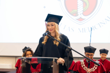 A person in a black academic gown and mortarboard speaks into a microphone at the lectern during an academic ceremony; people in red academic robes are seated in the background.