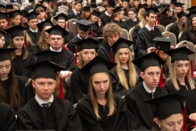 A large group of people in black academic gowns and mortarboards sit in rows during an academic ceremony.