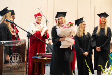 A person in a black academic gown and mortarboard holds a bouquet of flowers during an academic ceremony; other ceremony participants stand nearby.