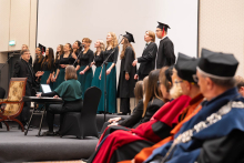 A choir performs on stage during an academic ceremony, accompanied by a keyboard instrument; people in ceremonial academic robes are seated in the foreground.