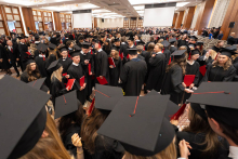 A large group of people in black academic gowns and mortarboards stand in a large hall after the academic ceremony; some are holding red folders.
