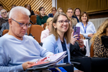 A man is looking through conference materials, while a woman seated next to him is holding a purple mobile phone. Conference participants are visible in the background.