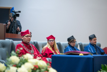 Four people in academic gowns sit in armchairs during the ceremony. A table with academic insignia stands in front of them, with a bouquet of white flowers in the foreground.