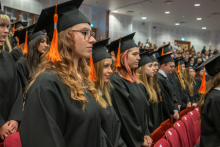 A group of graduates in black academic gowns and mortarboards with orange tassels stand in rows in the auditorium during the ceremony.