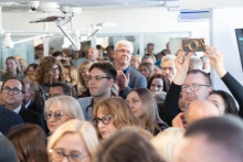 Participants fill the clinical room during the event. The faces of attendees can be seen in the foreground, while one person is recording the event on a mobile phone.