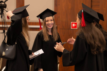 Three people in black academic gowns and mortarboards talk during an academic ceremony; one is holding a phone and an envelope.