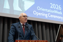The Rector speaks at the podium during the opening of Orientation Week 2026. A screen displaying the name of the event is visible in the background.