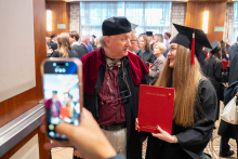 A person in a black academic gown and mortarboard poses with a red folder bearing the words “Medical Diploma” next to a person in red academic attire; someone is taking a photo with a phone in the foreground.