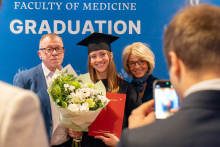 A person in a black academic gown and mortarboard poses with a bouquet of flowers and a red diploma folder against a backdrop bearing the words “Faculty of Medicine Graduation”; someone is taking a photo with a phone in the foreground.