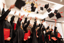 A group of people in black academic gowns toss their mortarboards into the air during a graduation ceremony; some are holding red diploma folders.