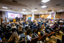Conference participants are seated in an auditorium listening to presentations taking place on stage.