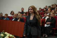 Three people in red academic gowns stand in front of a ceremony attendee wearing a light blue shirt. A person wearing a red mortarboard and a ceremonial chain shakes her hand while holding a white bag in the other hand. A list of names is displayed on a screen in the background.