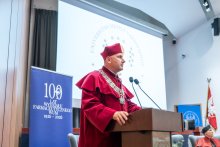 A person wearing a red academic gown and ceremonial chain speaks at a lectern. In the background, there is a banner marking the 100th anniversary of the WUM Faculty of Pharmacy.