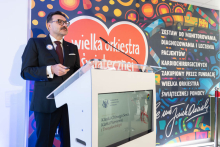 A person speaks at a lectern in a room at the Department of Cardiac, Thoracic Surgery and Transplantology. In the background, there is a WOŚP graphic and a sign indicating the purchase of a system for monitoring, diagnosing and treating cardiac surgery patients.