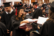 People in black academic gowns and mortarboards hand red folders to one another during an academic ceremony.
