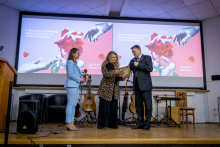 Two women and a man stand on stage during a ceremony. The woman on the left is holding a red rose, the woman in the centre is holding a framed certificate, and the man is holding a microphone.