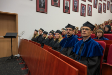 People wearing blue academic gowns and black caps sit in a row in an auditorium. Other ceremony participants are seated behind them.