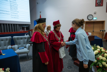 A person wearing a red academic gown and a black mortarboard speaks at a lectern with two microphones, holding an open folder. Behind the lectern stand three people in red academic gowns and mortarboards, two of them wearing ceremonial chains.