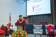 A person in an academic gown and mortarboard speaks at the lectern during the Graduation Ceremony of the Faculty of Medicine and Dentistry of the Medical University of Warsaw. The screen displays the name of the event and the date, 14 October 2026.