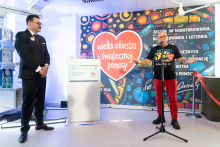 Two people stand in front of a WOŚP graphic in a clinical room. One person speaks into a microphone, while the other listens beside the lectern.