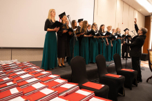 A choir performs on stage during an academic ceremony, led by a conductor. Stacks of red folders are placed in the foreground.