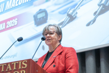 A woman speaks at a lectern equipped with two microphones. In the background, a presentation related to the Polish Championship in Minimally Invasive Surgery is displayed.