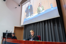 Mężczyzna w todze z łańcuchem sędziowskim siedzi przy stole w auli, podczas gdy na dużym ekranie za nim wyświetlana jest transmisja z dwiema osobami.