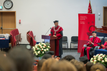 The Dean, wearing academic dress, delivers a speech at the lectern. A burgundy backdrop featuring the faculty logo and several people seated on stage are visible in the background.