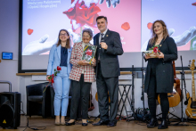 Two women, an older woman, and a man pose for a photo on stage. The women are holding red roses, and the older woman is also holding a framed artwork.