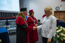 A person in a white shirt stands opposite a person wearing a red academic gown and a black mortarboard