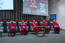 Five people in ceremonial academic gowns are seated on the stage in the auditorium. Behind them is a large screen showing a live feed from the lectern.