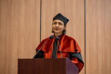 A person wearing a red-and-black academic gown, mortarboard, and ceremonial chain speaks at a lectern.