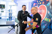 Two people shake hands by a microphone during a ceremony in a clinical room. A WOŚP graphic and medical equipment can be seen in the background.
