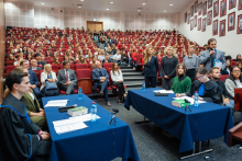 Szeroki plan auli uniwersyteckiej, w której odbywa się symulacja rozprawy sądowej; uczestnicy w todze i garniturach siedzą przy stołach, a na widowni zasiada tłum studentów.