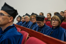 People wearing blue academic gowns and black caps sit in a row in an auditorium. Other ceremony participants are seated behind them.