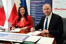 Dwie osoby siedzą przy stole i podpisują dokumenty. Na stole leżą dokumenty oraz plansza informująca o dofinansowaniu projektu. W tle widoczne są flagi Polski i Unii Europejskiej oraz materiały dotyczące Funduszy Europejskich dla Mazowsza.