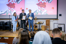 A woman and two men stand on stage during a ceremony. The woman is holding a red rose, and both men are holding framed documents. One of the men is speaking into a microphone.