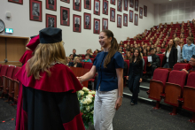 A person in a navy blue top and white trousers shakes hands with a person wearing a red academic gown and a black mortarboard. In the background, numerous ceremony attendees are seated or standing among rows of red chairs in the auditorium.