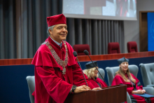 A person in a rector’s gown and mortarboard speaks at the lectern during the ceremony in the auditorium. A floral arrangement stands in front of the lectern.