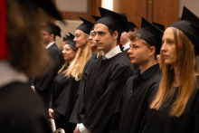 A group of people in black academic gowns and mortarboards stand in rows during an academic ceremony.