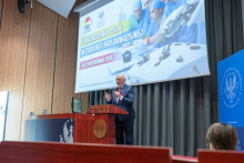 A man speaks at a lectern equipped with two microphones. In the background, a presentation on minimally invasive surgery is displayed.