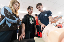 Three people watch a first aid demonstration using a manikin. The person conducting the demonstration is holding a medical device.