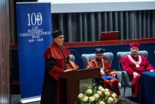Osoba w czerwono-czarnej todze akademickiej, birecie i łańcuchu ceremonialnym przemawia przy mównicy ozdobionej kompozycją z białych kwiatów.