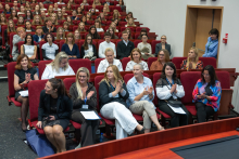 A group of people sit in the front rows of an auditorium during a ceremony, with some of them applauding. A large audience is seated in the rows behind them.