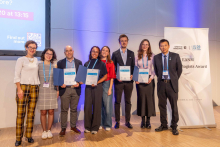 Siedem osób stoi na scenie podczas ceremonii wręczenia nagród EANM Technologists Award. Cztery osoby trzymają otwarte dyplomy. Po prawej stronie znajduje się roll-up z nazwą nagrody i logotypami wydarzenia. W tle widać ekran prezentacyjny oraz oświetlenie sceniczne.