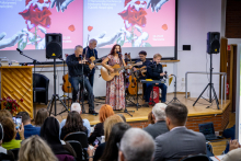 A music band performs on stage in front of an audience. A woman is singing and playing an acoustic guitar while the other musicians are playing instruments.