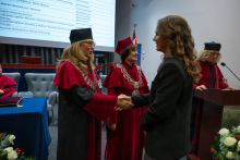 A person in a black suit shakes hands with a person wearing a red academic gown, a black mortarboard and a ceremonial chain. Next to them stands a person wearing a red gown, a red mortarboard and a ceremonial chain, while another person in an academic gown reads from a text at the lectern. A list of degree programmes and names is displayed on a screen in the background.